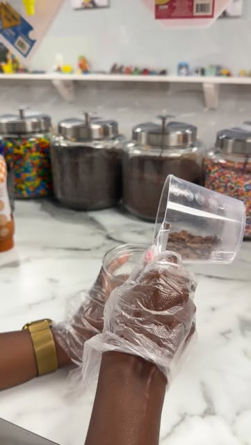 A Shaker being made at the counter, cereal poured into the cup in front of jars of sprinkles, cookie crumbs and cocoa
