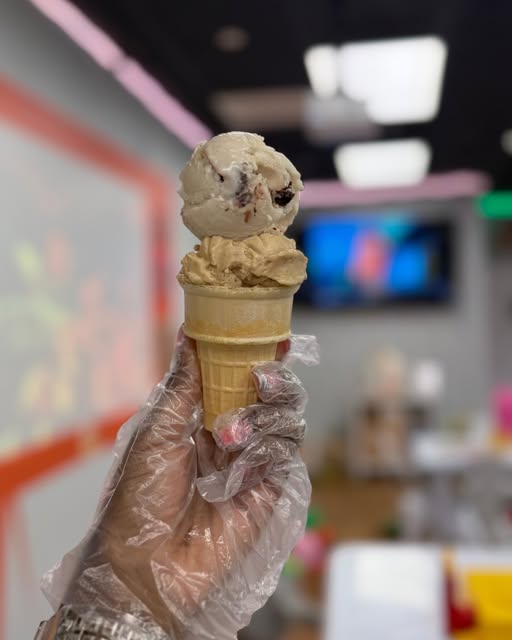 A two-scoop cone held up inside the shop, with the bright seating area and display screen behind it